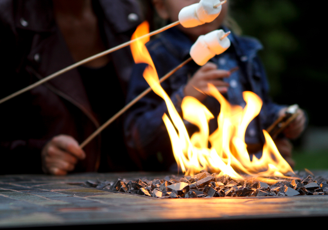 Two people roasting marshmallows over a propane firepit outside.